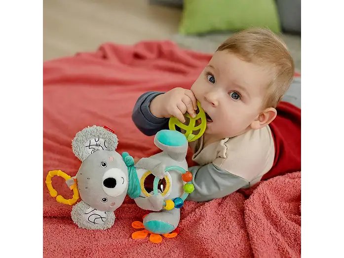 Dziecko trzyma zielony element zabawki FEHN DoBabyDoo Koala przy ustach, leżąc na czerwonym kocu.